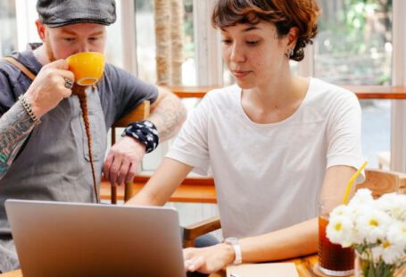 Remote Teams - Pensive young woman typing on netbook while man with cup drinking coffee at table of cafe